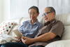 an elderly man and woman sitting on a couch looking at a tablet